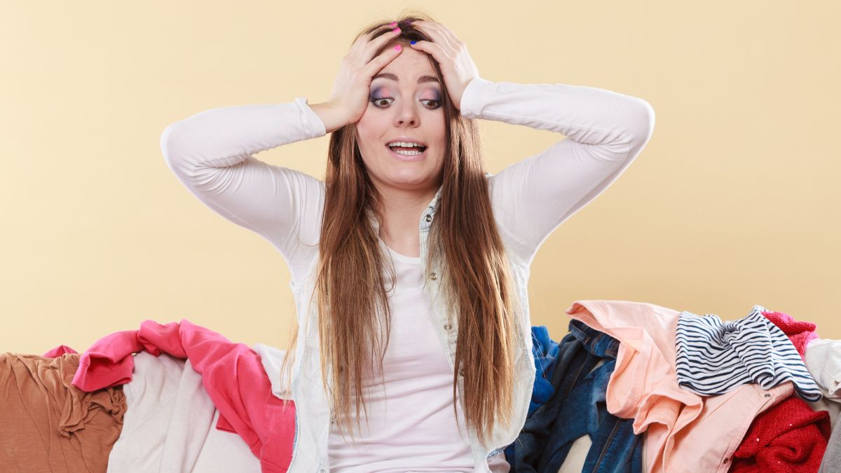 Woman with her hands on hear head overwhelmed with clothing all around her on the couch.